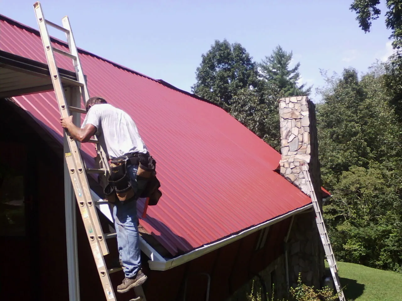 Roofer on a ladder beside a red metal roof and stone chimney — Soffit & Fascia Repair by Roofing Brothers in Nappanee